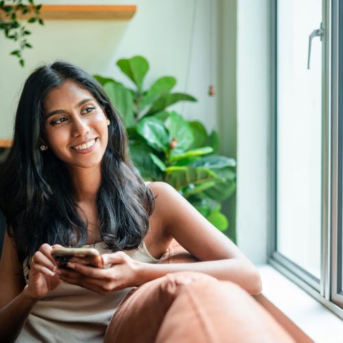 young Asian woman relaxing on sofa at home, using smartphone and looking away with smile