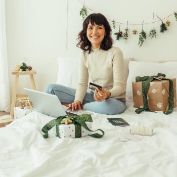 Happy woman sitting on bed in a decorated room with christmas presents doing online shopping. Woman using credit card for online shopping on laptop computer.